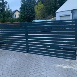 PHU SUN-TEL Cezary Dobrzyński - Nowoczesna, pozioma brama wjazdowa z automatyką, pomalowana na grafitowo, na tle kostki brukowej i zabudowań. Widoczne siłowniki i skrzynka sterująca.