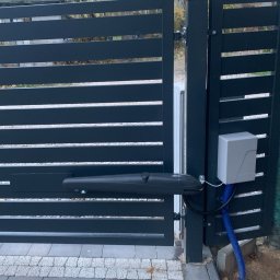PHU SUN-TEL Cezary Dobrzyński - Nowoczesna brama wjazdowa z automatyką: szare, poziome panele i siłownik. Widoczny sterownik i niebieski peszel. Kostka brukowa u podstawy.