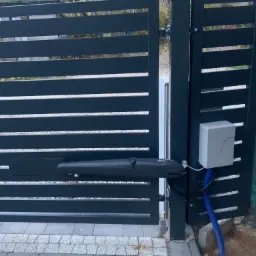Nowoczesna brama wjazdowa z automatyką: szare, poziome panele i siłownik. Widoczny sterownik i niebieski peszel. Kostka brukowa u podstawy.