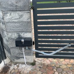 PHU SUN-TEL - Szara brama wjazdowa z automatyką, widoczny siłownik marki Beninca zamontowany na kamiennym słupku. Tło z kostki brukowej.