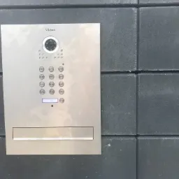 Panel wideodomofonu Vidos z kamerą i klawiaturą numeryczną, zamontowany na szarej, betonowej ścianie z bloków. Widoczne delikatne refleksy światła na metalowej obudowie.