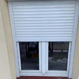 Zamknięta, biała roleta antywłamaniowa na oknie z widoczną fakturą tynku na ścianie budynku.