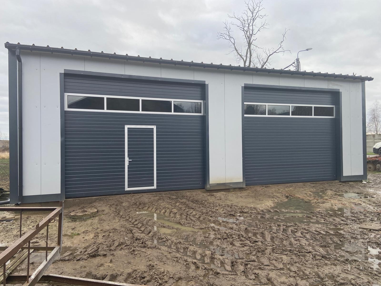 Dwie szare segmentowe bramy garażowe z oknami, umieszczone w białym budynku z metalową konstrukcją, widoczne ślady opon na błotnistym podłożu.