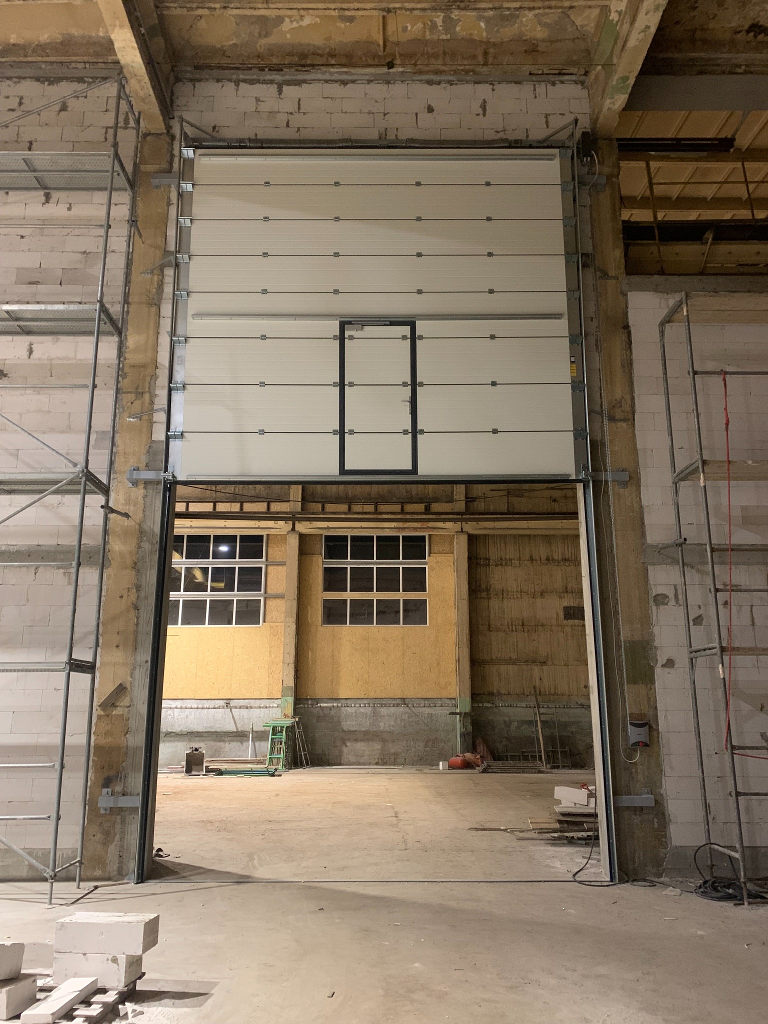 Biała segmentowa brama garażowa z wbudowanymi drzwiami, widok od wewnątrz hali w trakcie montażu, rusztowania po bokach, surowe ściany i podłoga.