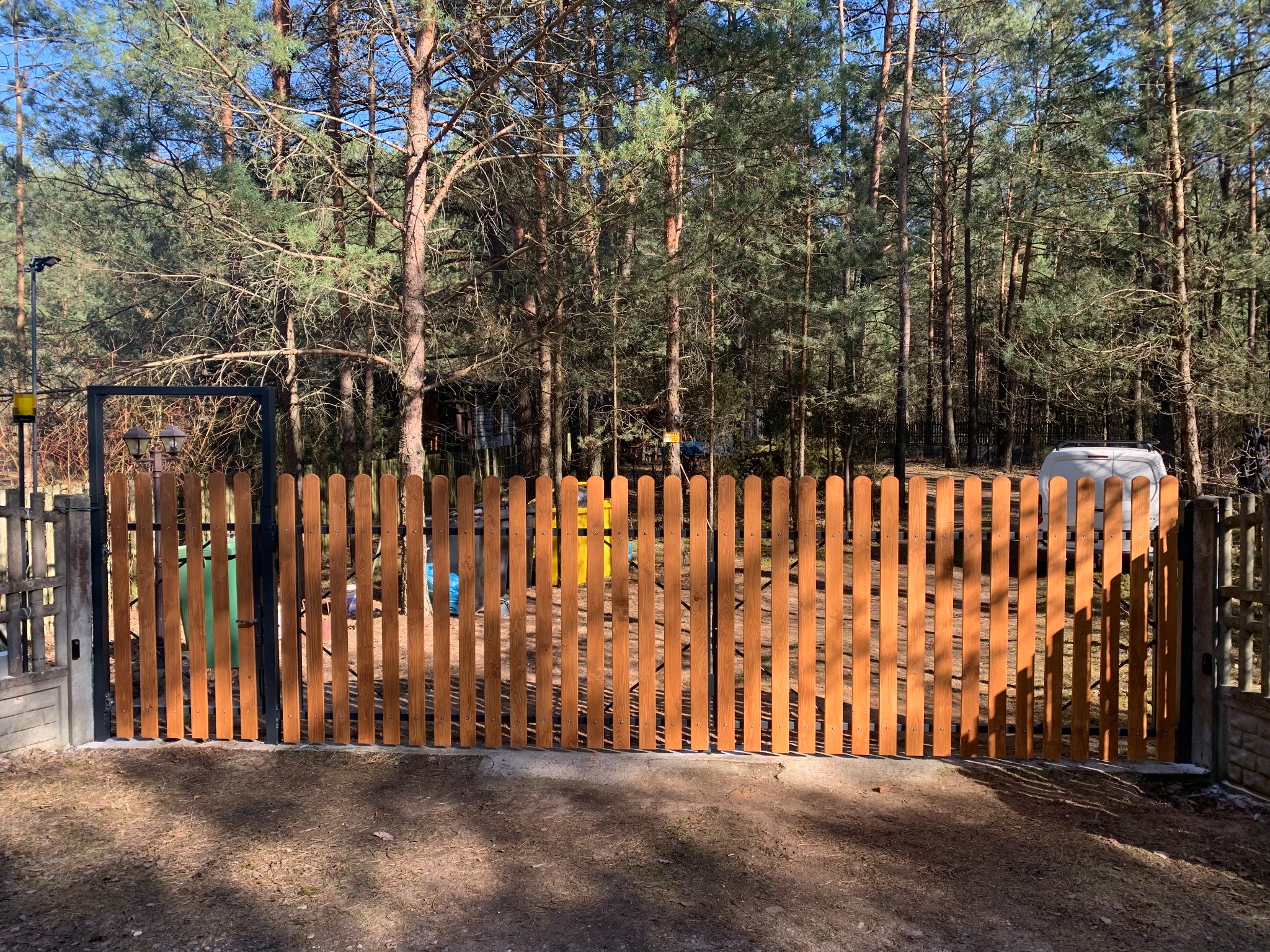 Nowe drewniane ogrodzenie z furtką, widok z zewnątrz, w tle las iglasty i zaparkowany biały samochód dostawczy.