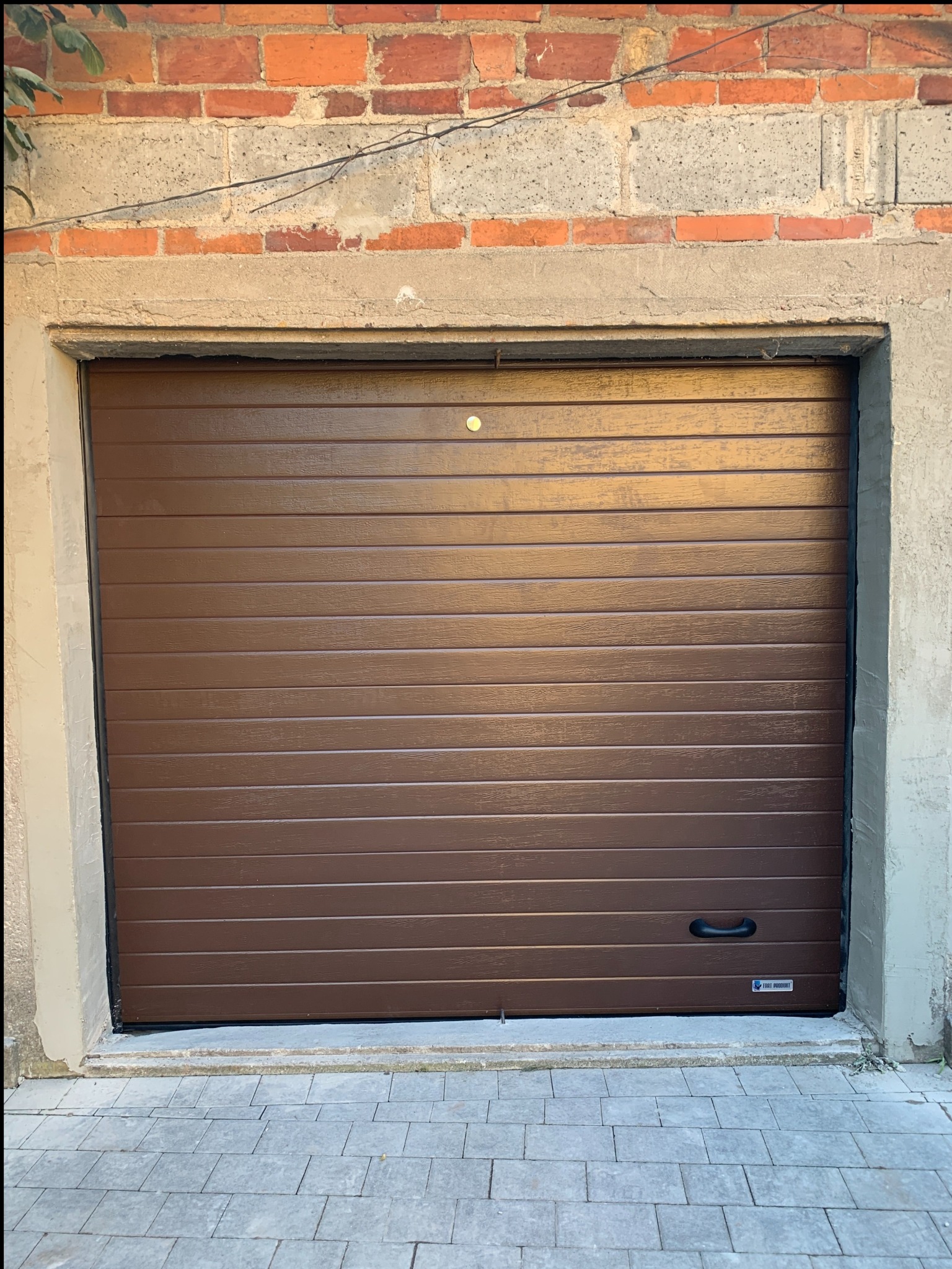 Zamknięta, brązowa brama garażowa segmentowa z poziomymi przetłoczeniami, zamontowana w surowym, ceglano-betonowym otworze. Widoczny fragment brukowanej podłogi.