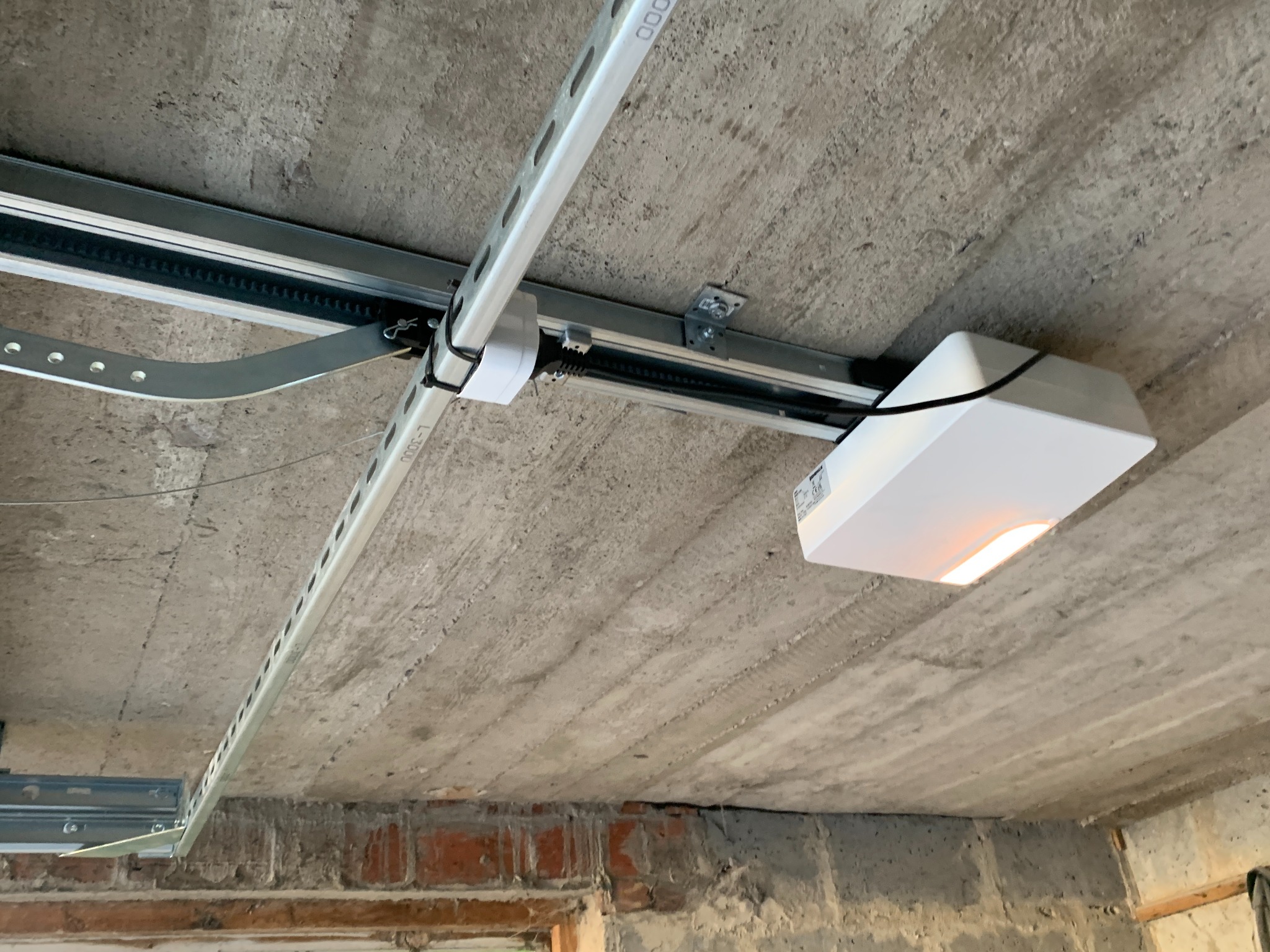 Zamontowany napęd bramy garażowej pod surowym betonowym stropem, widoczne prowadnice i elementy mocujące. Oświetlenie napędu włączone.