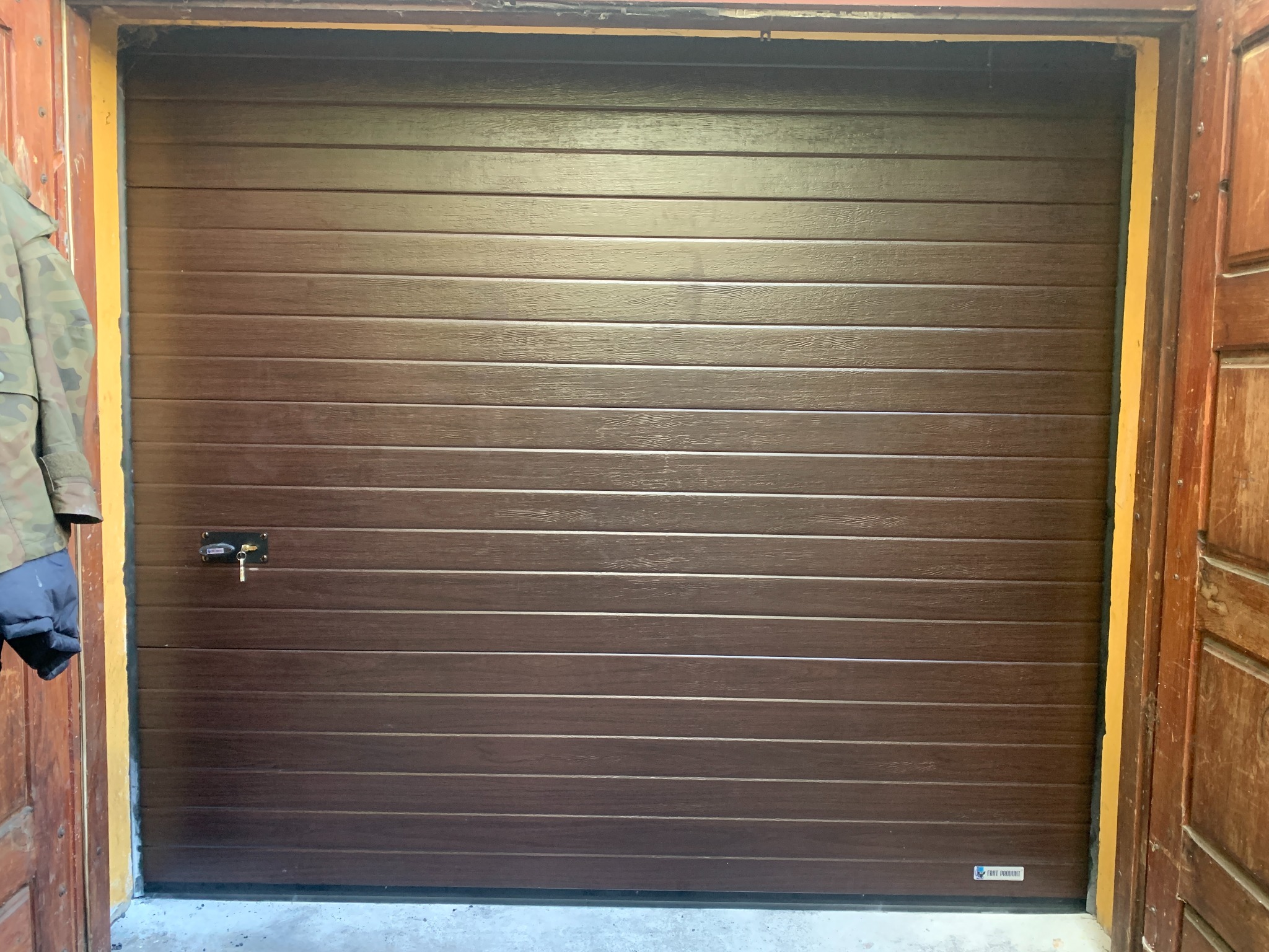 Zamknięta, segmentowa brama garażowa w kolorze brązowym z widocznym zamkiem i kluczem, zamontowana w drewnianej ościeżnicy. Po lewej fragment kurtki.