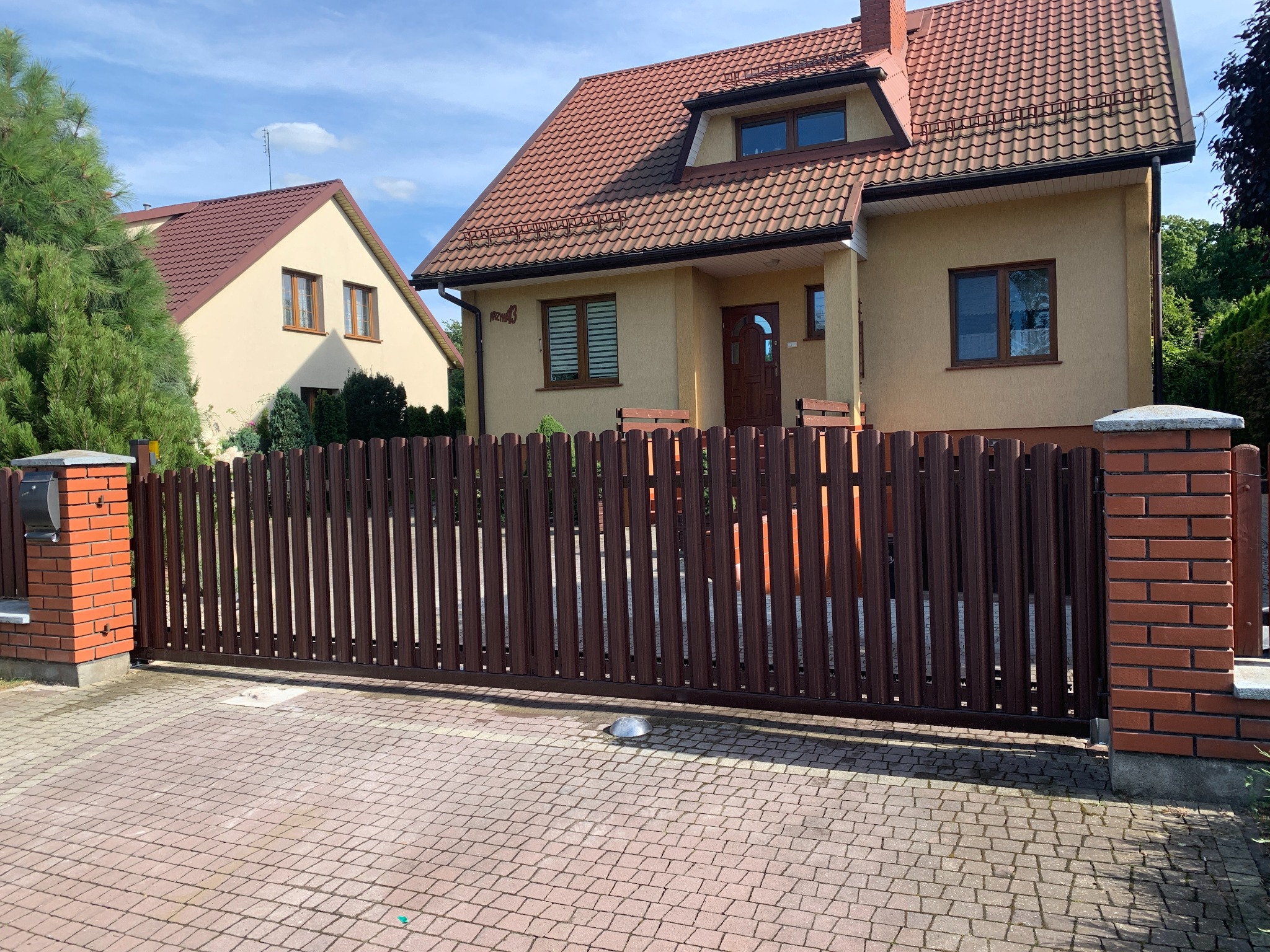 Brązowa, przesuwna brama wjazdowa z pionowych sztachet, zamontowana na posesji z dwoma domami o beżowych elewacjach i brązowych dachach, widok z zewnątrz.