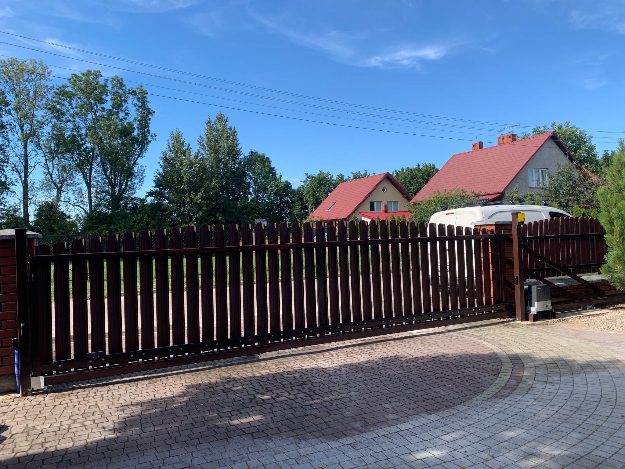 Automatyczna brama przesuwna z brązowych sztachet, z widocznym napędem, na tle domów z czerwonymi dachami i błękitnego nieba.