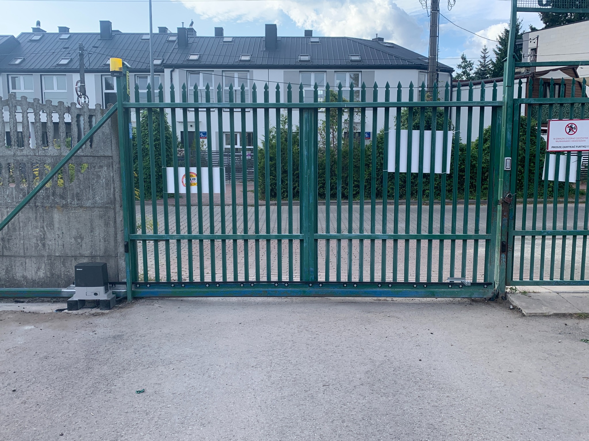 Zielona brama wjazdowa z automatyką na tle budynku. Widoczny napęd bramy marki Sommer. Słupki bramy z tabliczkami i ostrzeżeniem.
