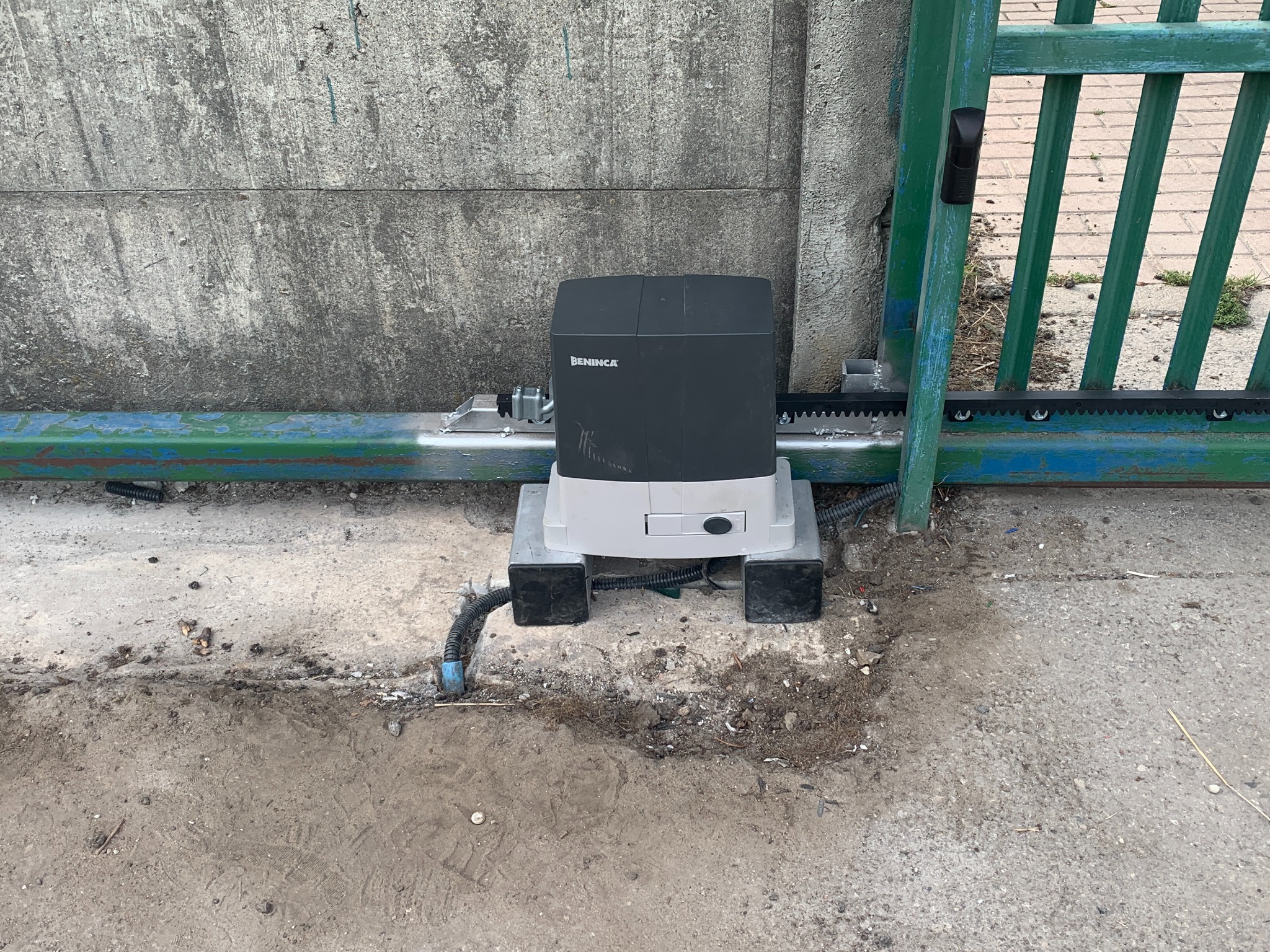 Zamontowany automat BENINCA przy bramie przesuwnej z zębatą listwą. Widoczne przewody i podstawa automatu na tle betonowego podłoża i fragmentu zielonej bramy.