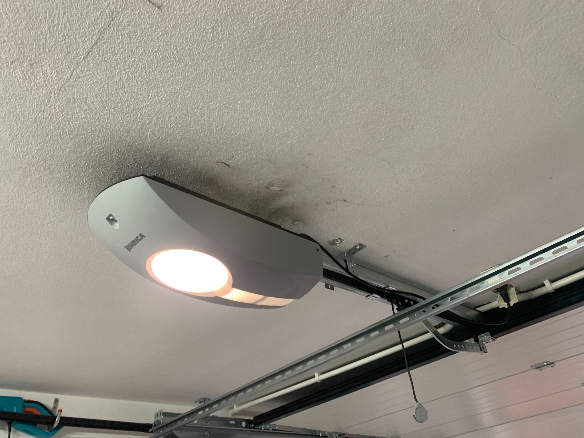 Zamontowany automat do bramy garażowej marki Beninca, widoczny od dołu, z włączonym oświetleniem, przymocowany do sufitu garażu, z widocznymi elementami konstrukcyjnymi bramy.