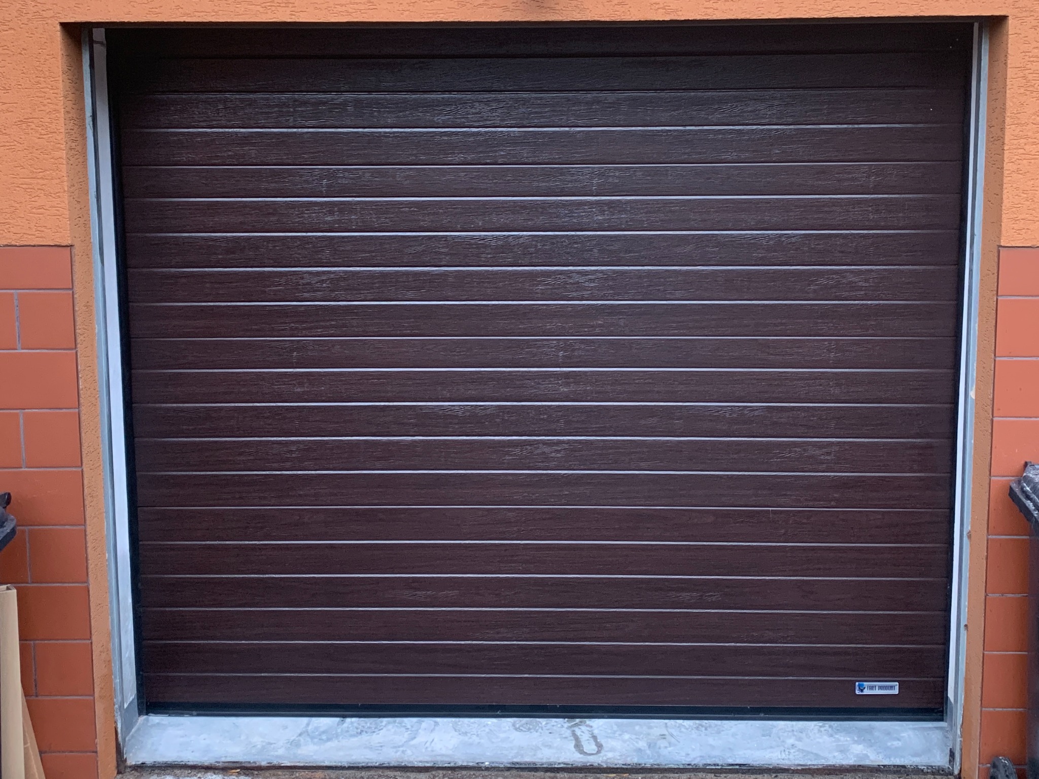 Zamknięta brama garażowa segmentowa w kolorze ciemnego drewna, z poziomymi przetłoczeniami, zamontowana w pomarańczowym budynku. Widoczny fragment muru z cegły.