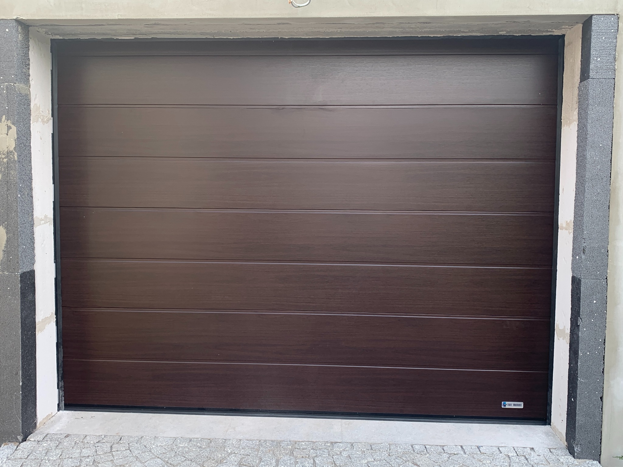 Ciemnobrązowa segmentowa brama garażowa z poziomymi panelami o strukturze drewna, zamontowana w surowym otworze garażowym z widocznym ociepleniem.