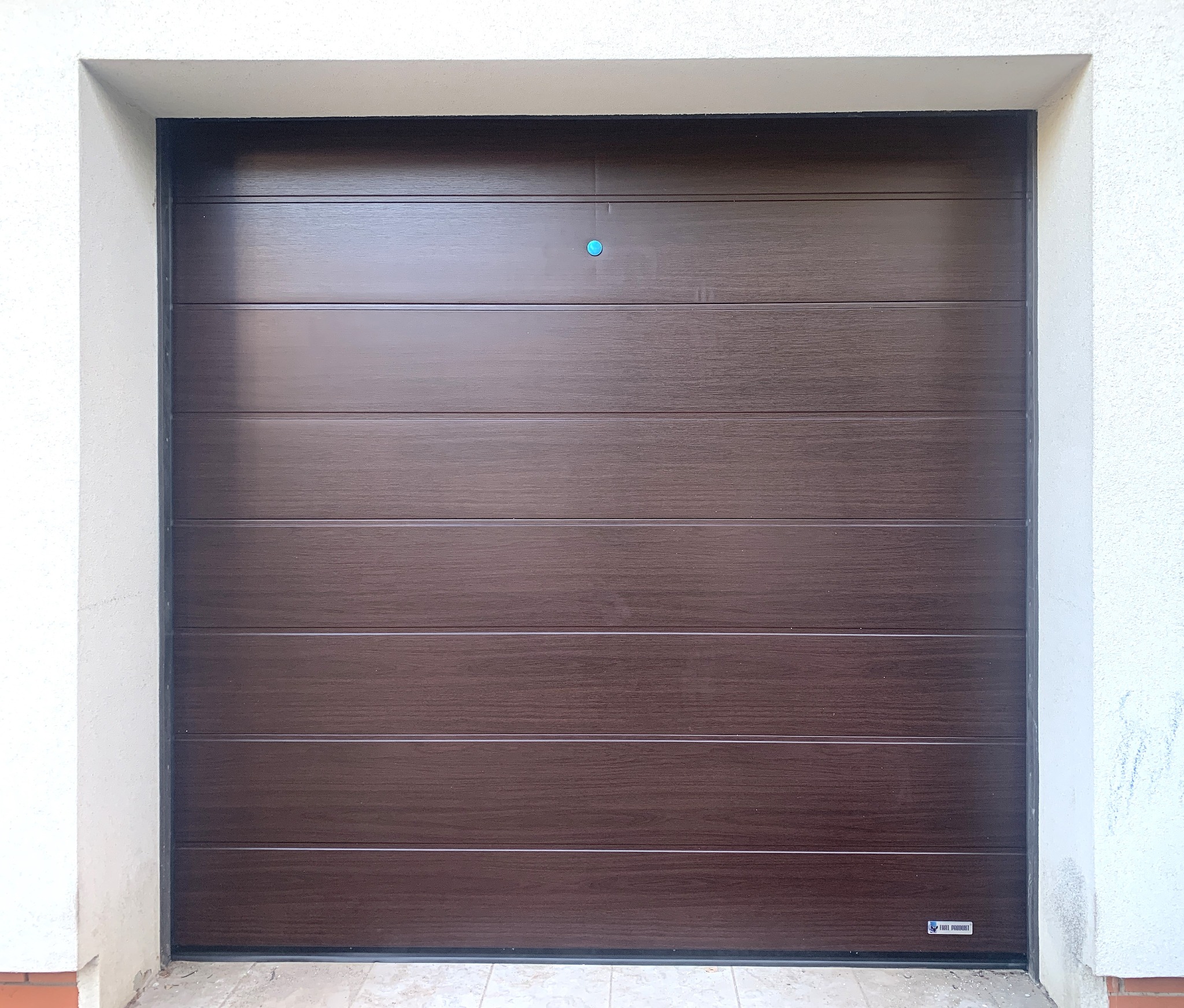 Zamknięta, segmentowa brama garażowa w odcieniach brązu z delikatną fakturą drewna, widoczna od frontu, z niebieskim punktem pośrodku i logo producenta w prawym dolnym rogu.