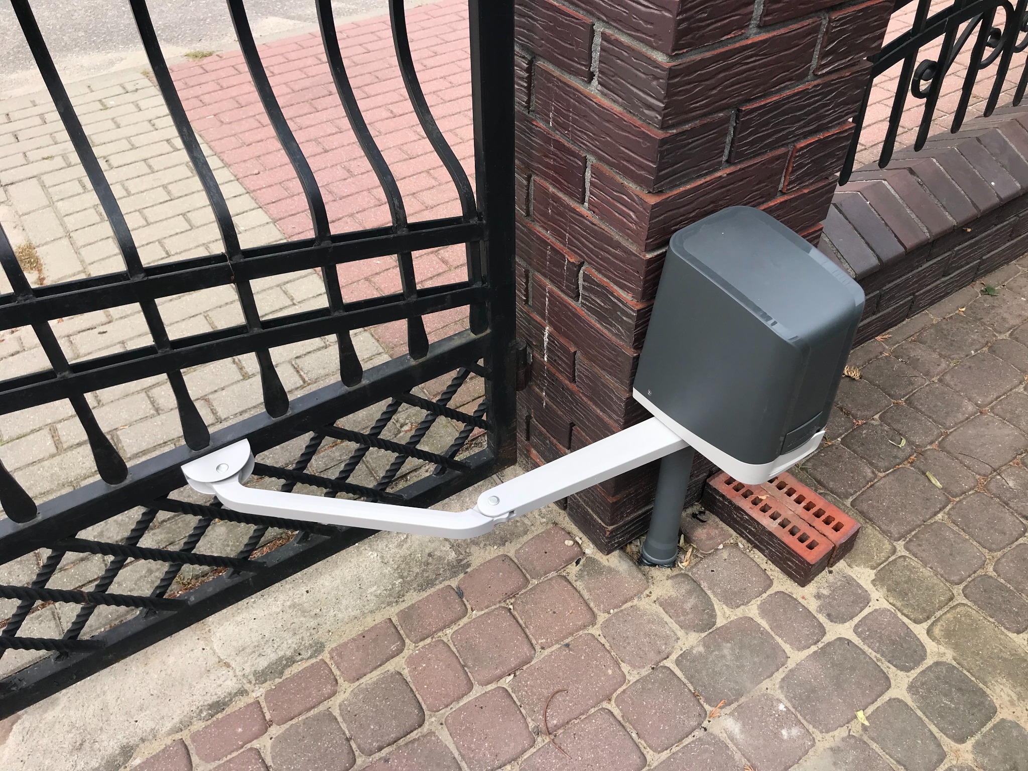 Czarna, metalowa brama wjazdowa z zamontowaną automatyką (szary napęd z białym ramieniem) przy słupku z czerwonej cegły, na tle kostki brukowej.