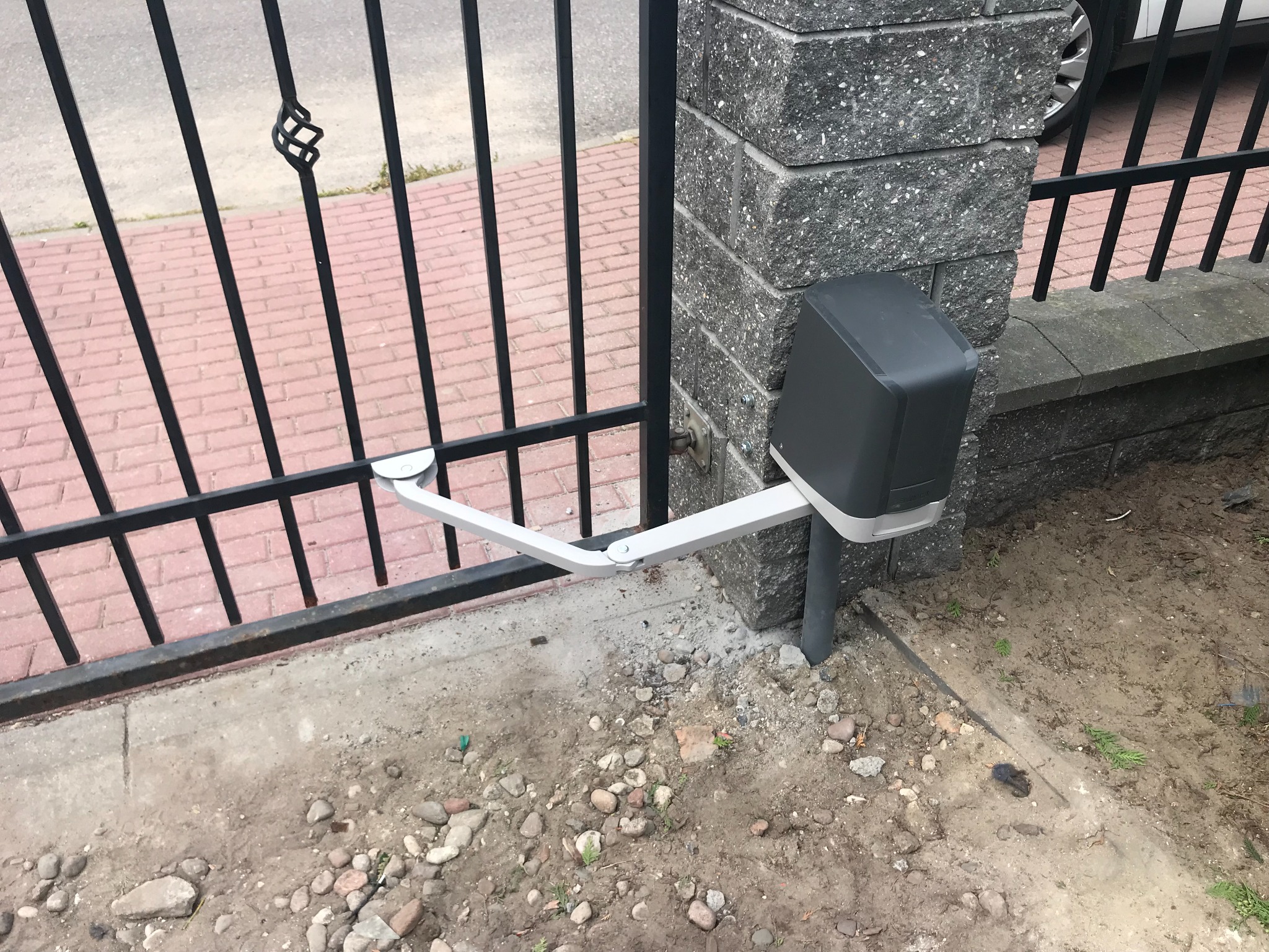 Szara automatyka do bramy skrzydłowej, zamontowana na murowanym słupku ogrodzeniowym. Widoczna czarna brama z ozdobnym elementem i fragment chodnika z czerwonej kostki.