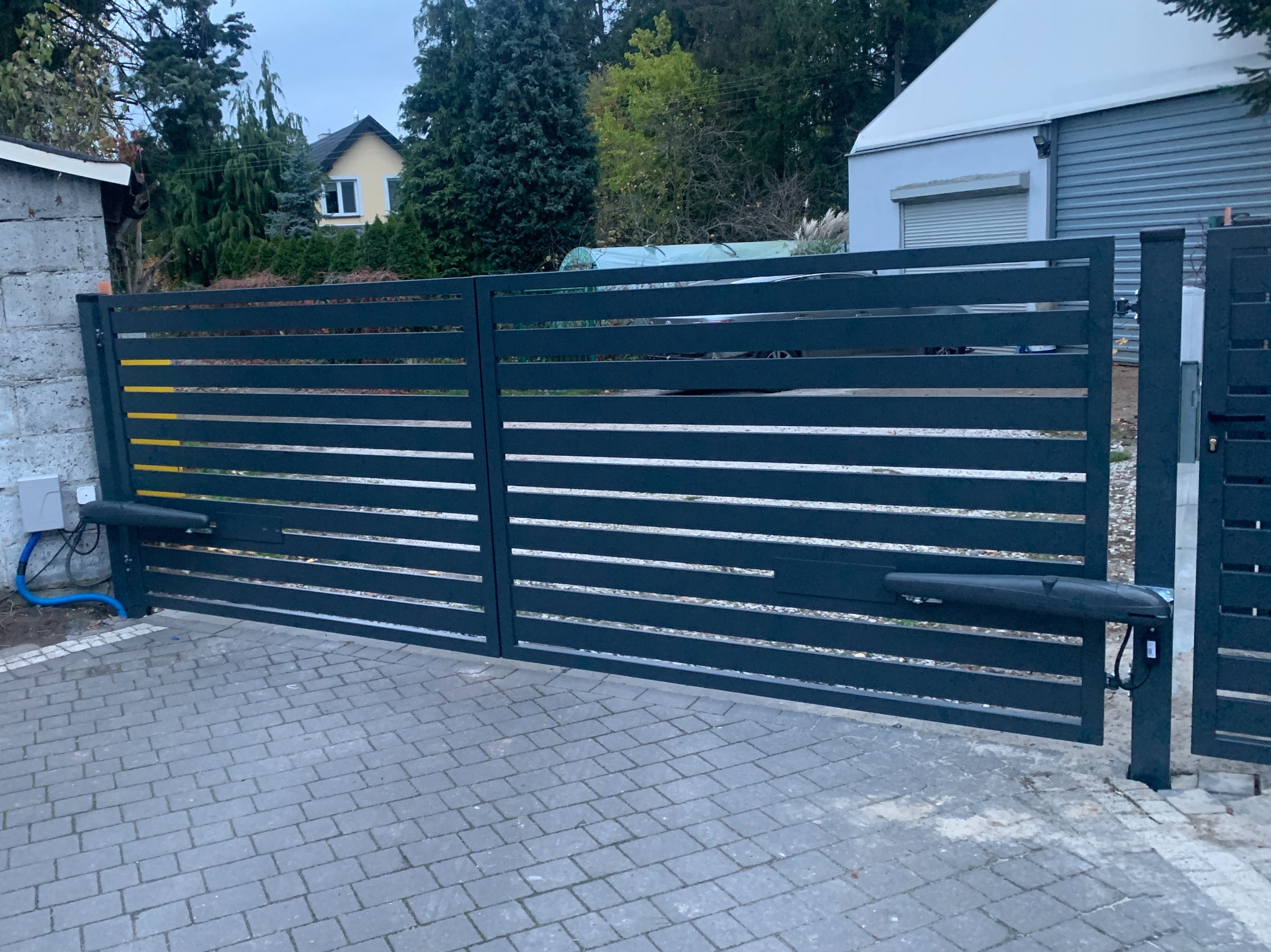 Nowoczesna, pozioma brama wjazdowa z automatyką, pomalowana na grafitowo, na tle kostki brukowej i zabudowań. Widoczne siłowniki i skrzynka sterująca.