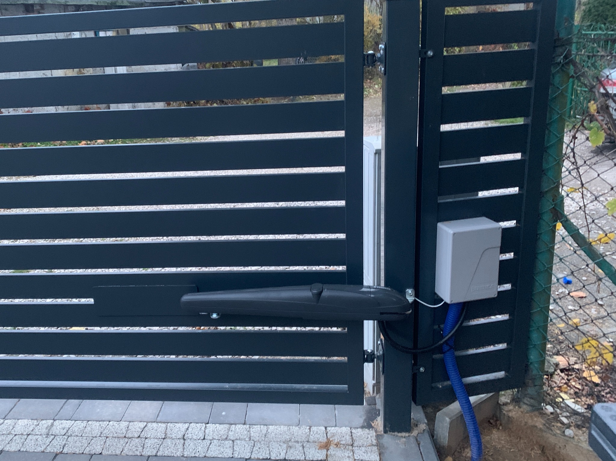 Nowoczesna brama wjazdowa z automatyką: szare, poziome panele i siłownik. Widoczny sterownik i niebieski peszel. Kostka brukowa u podstawy.