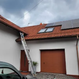 Dom z czerwoną dachówką i panelami słonecznymi, drabina oparta o ścianę. Widoczne okna dachowe i brama garażowa. Częściowo widoczny samochód.