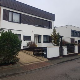 Kos-Mal Malowanie dachów i elewacji - Biały dom z brązowymi elementami drewnianymi i numerem 10. Widoczny fragment kostki brukowej i ogrodzenia. Drugi budynek w tle.