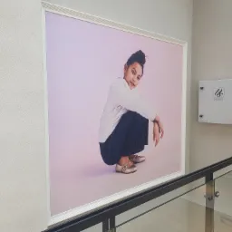 Duży, oprawiony plakat reklamowy z młodą dziewczyną w kucki na różowym tle, widoczny zza szklanej barierki na korytarzu.