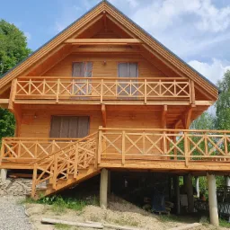 Nowo wybudowany, dwukondygnacyjny drewniany dom z balkonami, jeszcze w trakcie prac wykończeniowych, z widocznymi foliami ochronnymi na oknach, otoczony zielenią i elementami budowlanymi.