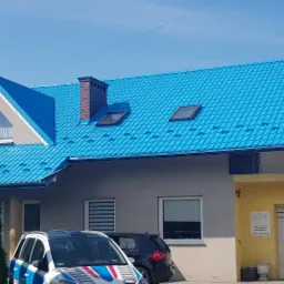 Dom jednorodzinny z nowym, błyszczącym dachem z blachodachówki w kolorze turkusowym, widoczne okna dachowe i komin z cegły, na pierwszym planie zaparkowane samochody.