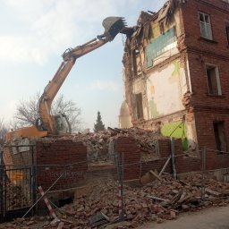 USB - Żółta koparka z wysięgnikiem burzy ceglany, częściowo zniszczony budynek z pozostałościami okien i fragmentami płytek, zabezpieczony siatką, na tle pochmurnego nieba.