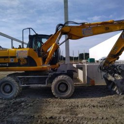USB - Żółta koparka kołowa JCB JS145 na placu budowy, obok wylany fundament pod budynek, widoczne zbrojenia i elementy konstrukcyjne.