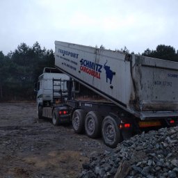 USB - Ciężarówka Schmitz z podniesioną naczepą, wysypująca gruz na błotnistej działce, w tle las iglasty i pochmurne niebo.