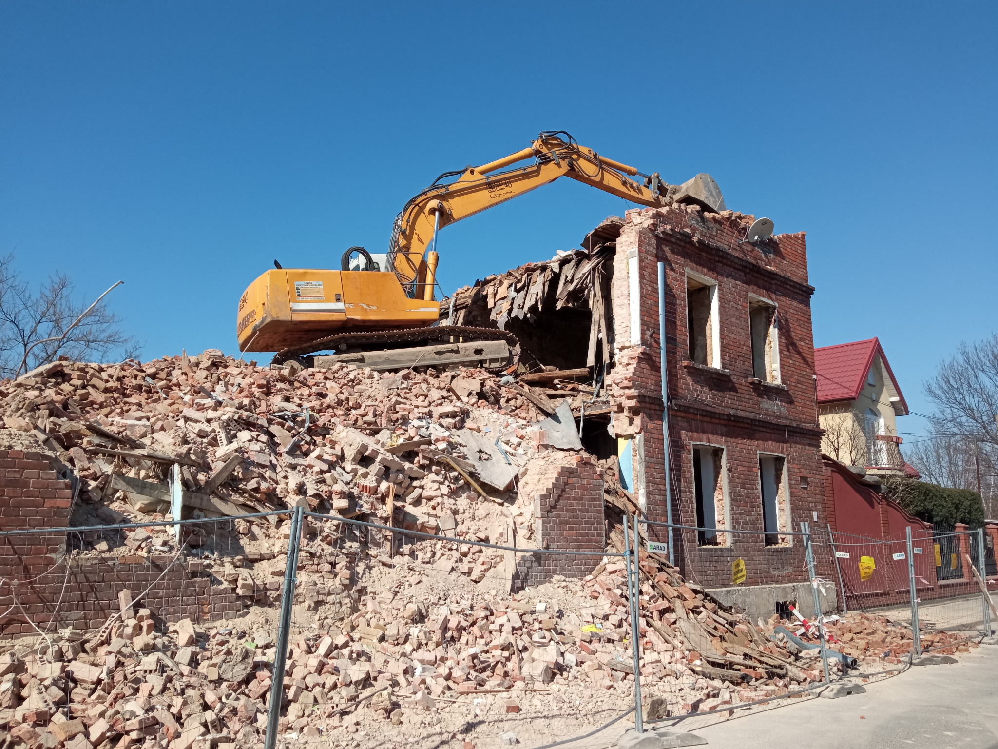 Żółta koparka na szczycie częściowo zburzonego, ceglanego budynku z widocznymi fragmentami ścian i stertą gruzu; w tle błękitne niebo i fragment budynku z czerwonym dachem.