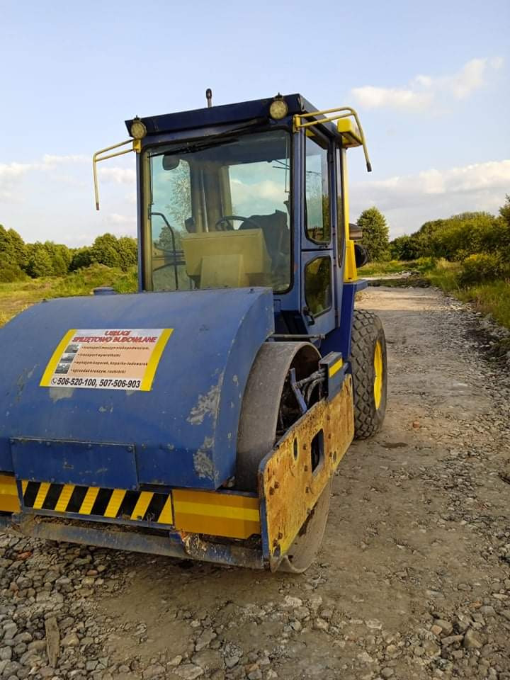 Niebiesko-żółty walec drogowy z widocznymi śladami użytkowania stoi na żwirowej drodze, z naklejką reklamową na boku i drzewami w tle.