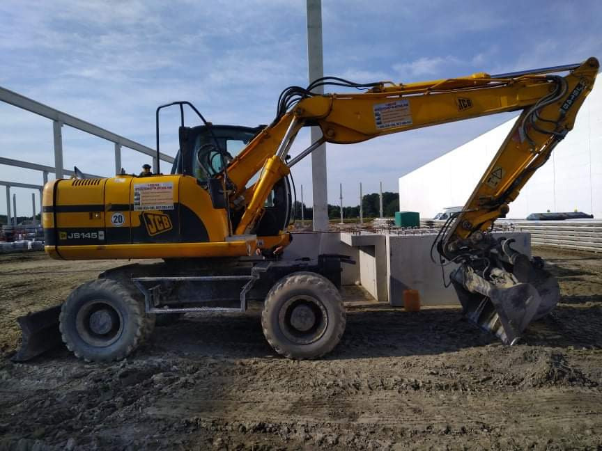 Żółta koparka kołowa JCB JS145 na placu budowy, obok wylany fundament pod budynek, widoczne zbrojenia i elementy konstrukcyjne.