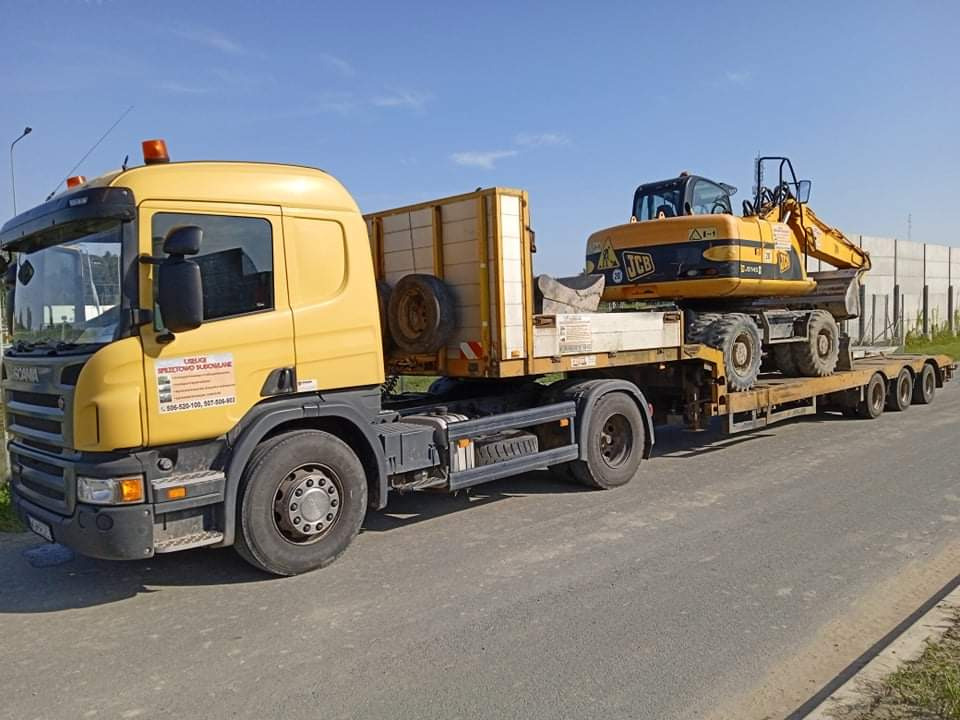 Żółta ciężarówka Scania z lawetą przewożąca żółtą koparkę JCB po asfaltowej drodze, widoczny znak firmy na kabinie ciężarówki.