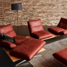 Nowoczesne, skórzane leżanki w odcieniach czerwieni na drewnianych podstawach, ustawione na dywanie w salonie z kamienną ścianą i lampą podłogową.