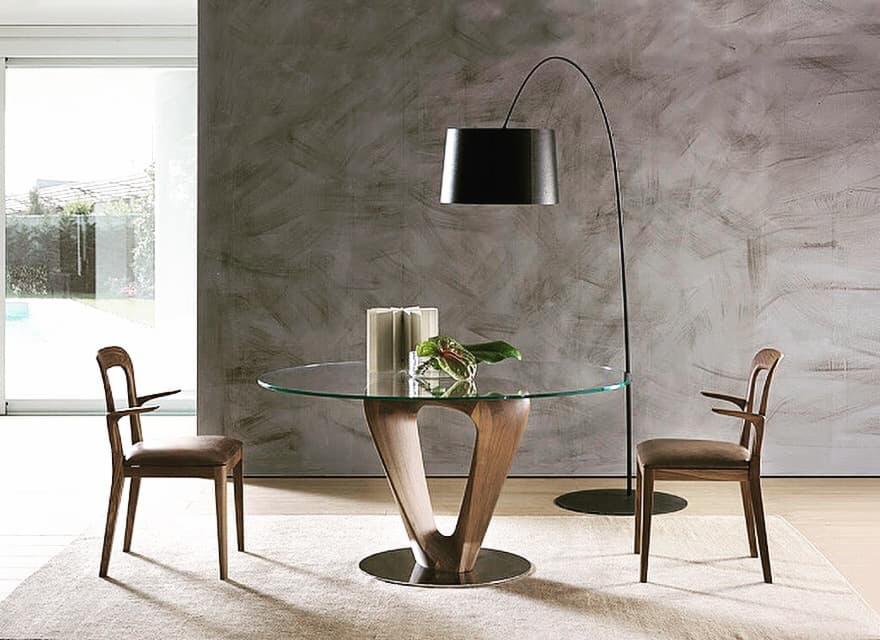 Okrągły stół z blatem ze szkła i drewnianą podstawą w kształcie litery V, otoczony dwoma drewnianymi krzesłami z podłokietnikami, oświetlony lampą podłogową z czarnym abażurem na tle ściany...