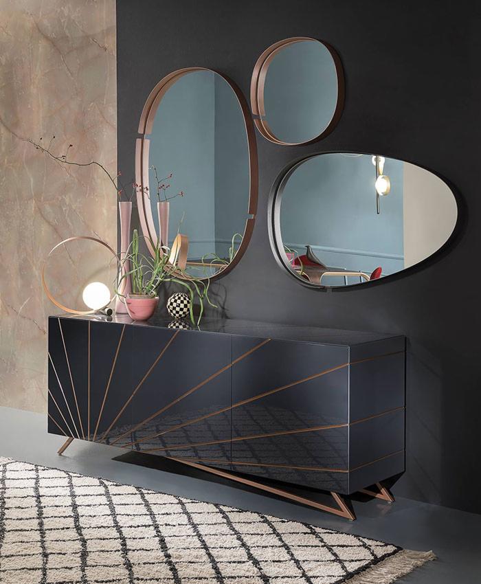 Czarny, lakierowany kredens z geometrycznym wzorem w kolorze miedzi, uzupełniony o kompozycję z trzech luster o różnych kształtach i okrągłej lampy, tworzący nowoczesną aranżację wnętrza.
