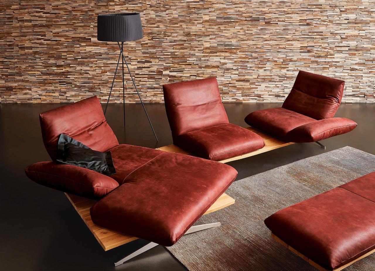 Nowoczesne, skórzane leżanki w odcieniach czerwieni na drewnianych podstawach, ustawione na dywanie w salonie z kamienną ścianą i lampą podłogową.