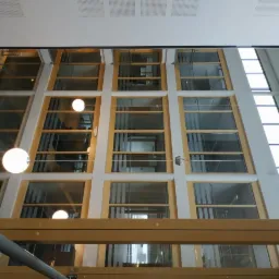 Wnętrze biurowca z wysokimi oknami i białymi ścianami z delikatnym kremowym odcieniem, widok z dołu na górę, z widocznymi lampami kulistymi i elementami konstrukcyjnymi sufitu.