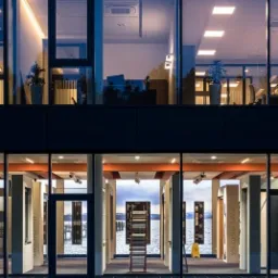 Wystawa różnorodnych drzwi w salonie z widokiem na jezioro, widoczny dwupoziomowy budynek z dużymi oknami, w tle biurowe wnętrze.