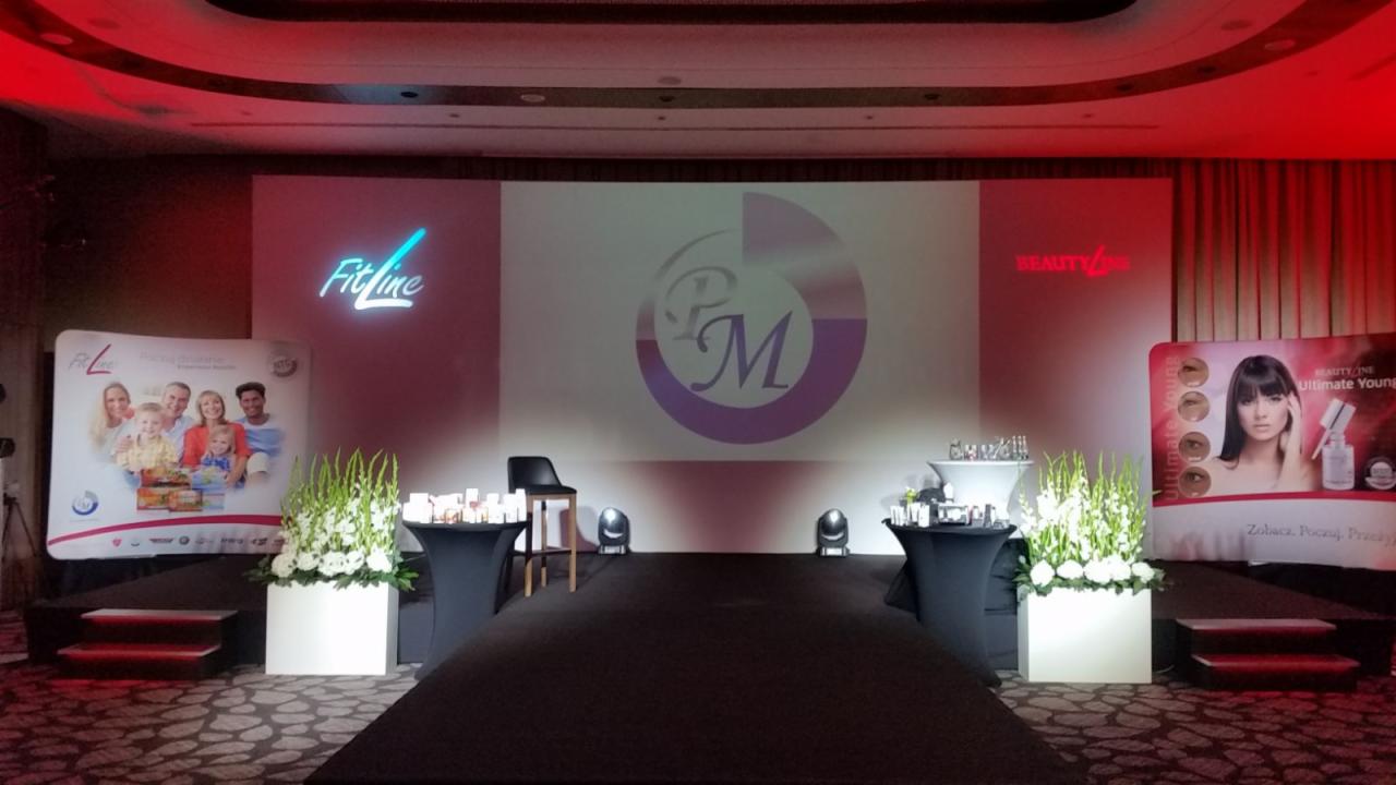 Scena z podświetlonym logo Fitline i Beautyline, dekoracje kwiatowe w białych donicach, czarny dywan i stoliki z kosmetykami, przygotowana do prezentacji produktów.