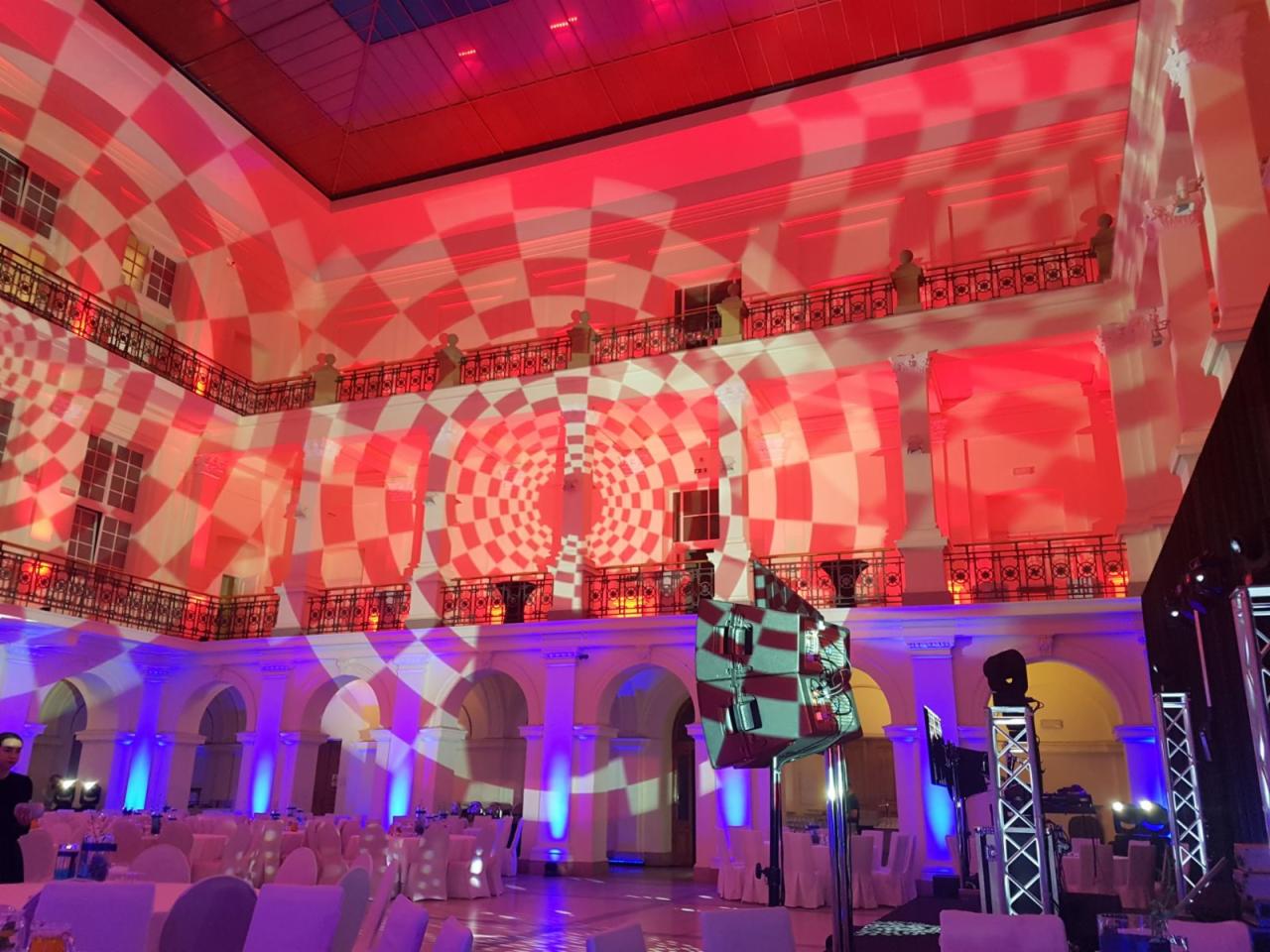 Wnętrze sali balowej z dekoracyjnym oświetleniem w czerwono-białą szachownicę wyświetlaną na ścianach i suficie, niebieskie reflektory oświetlają podłogę i ustawione stoły z krzesłami.