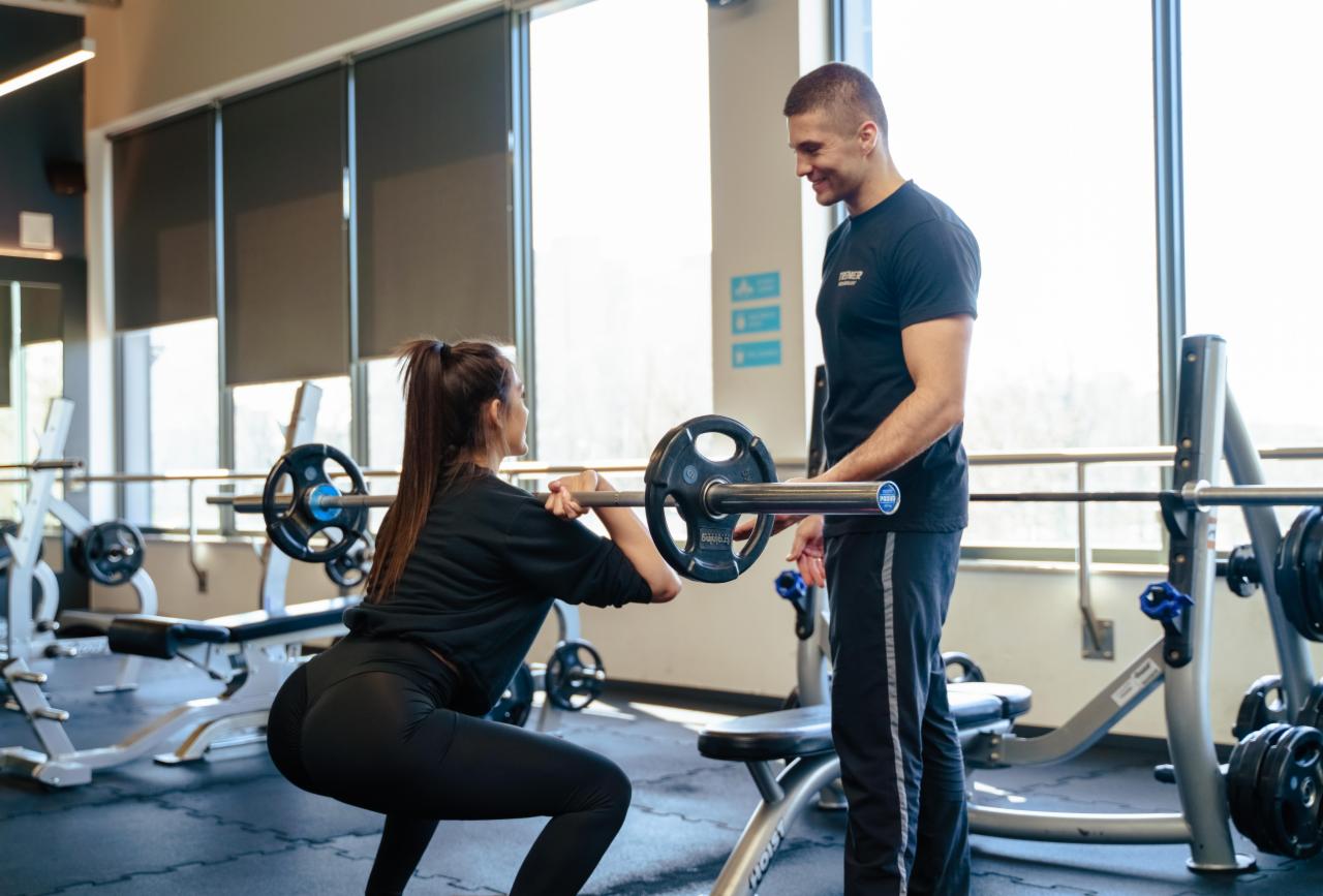 Kobieta wykonuje przysiad ze sztangą pod okiem trenera w jasnej siłowni z dużymi oknami.