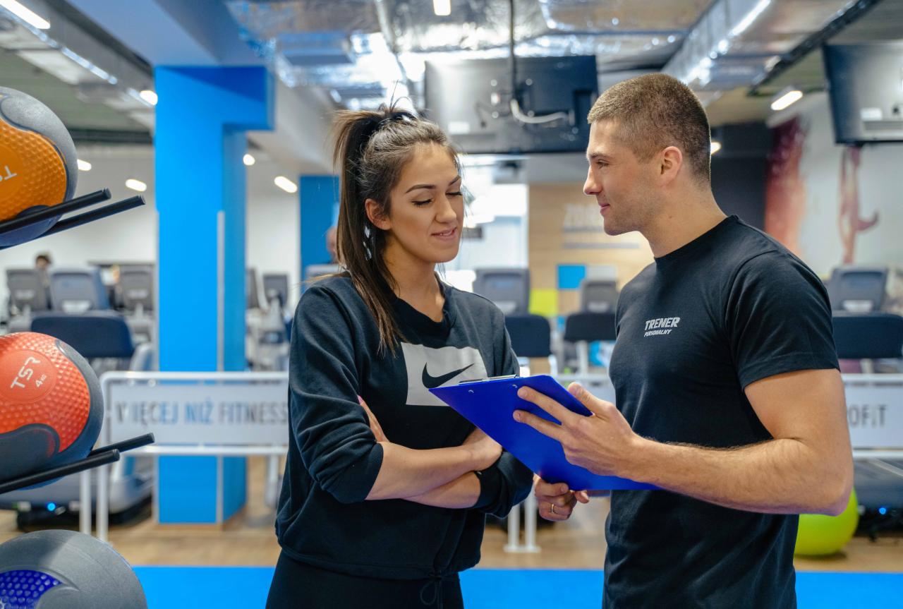 Trener personalny omawia plan treningowy z klientką w siłowni, na tle sprzęt do ćwiczeń i piłki lekarskie.