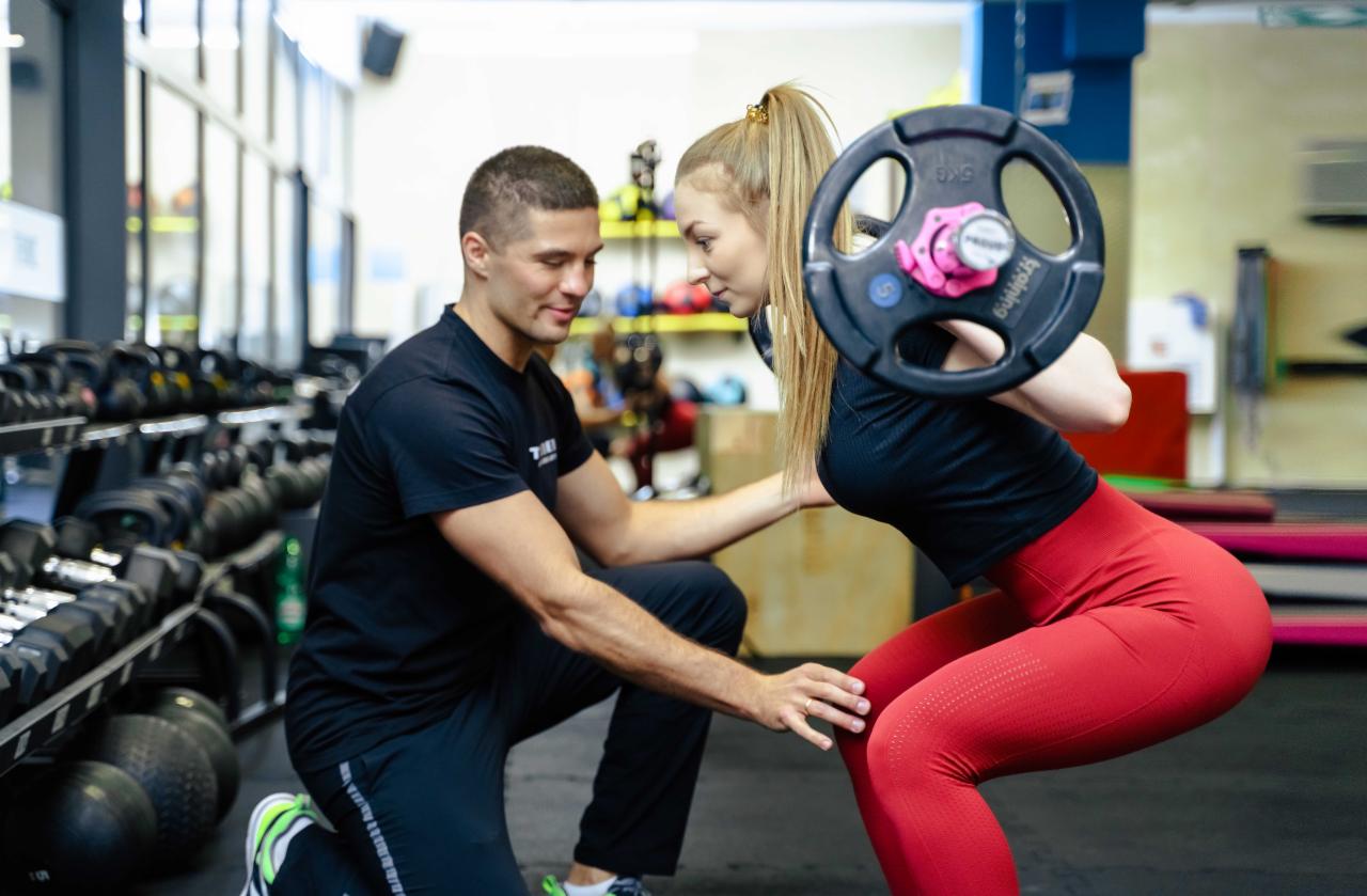 Trener personalny asekuruje młodą kobietę wykonującą przysiad ze sztangą w siłowni, widoczne obciążenia z różowymi zaciskami.