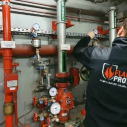 Pracownik w uniformie z logo firmy FLAME PROTECT reguluje zawór w systemie tryskaczowym przeciwpożarowym.