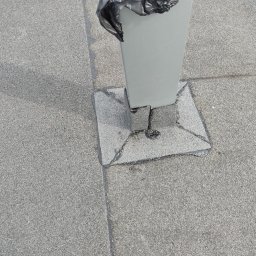 Usługi budowlane klimek - Szary, płaski dach pokryty papą z widocznym słupkiem zabezpieczonym czarną folią, uszczelnionym wokół podstawy.