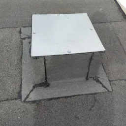 Szczelne przejście wentylacyjne na dachu pokrytym papą, zabezpieczone metalową osłoną z czterema śrubami.